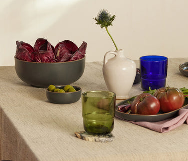 A table set with bowls of radicchio, olives, tomatoes, dates, and shredded red vegetables, along with crispbread, cheese, green glasses, and decorative white vases on a beige tablecloth.