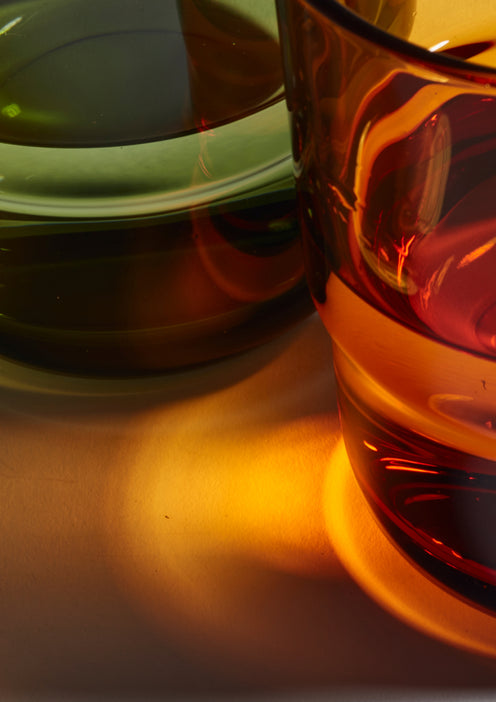 Close-up of two glass cups, one green and one amber, casting colorful reflections and shadows on a white surface in warm light.