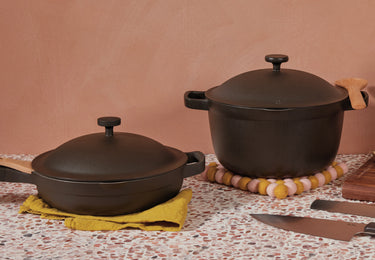 Two black cast iron pots with lids are on a terrazzo countertop; one is a deep pot on a felt trivet, and the other is a shallow pan on a yellow cloth. Two kitchen knives lie nearby against a peach-colored wall.