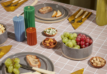 A table set with gray plates holding grapes, bread, and cheese, surrounded by bowls of mixed nuts and grapes. Three ribbed candles—green, blue, and orange—sit in the center. Yellow napkins and cutlery are neatly arranged.