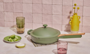 Translation missing: en.A green nonstick skillet with a lid and wooden handle sits on a mat atop a light countertop. Nearby are a plate of lime wedges, a small bowl, stacked glasses, a pink glass, and a yellow vase with flowers