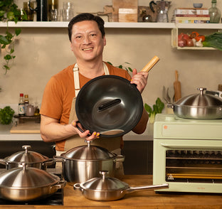 Man holding a black wok in a kitchen with various cookware and appliances.