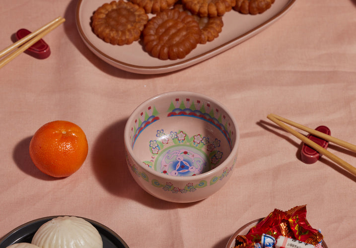 A decorative bowl with floral patterns sits on a pink cloth, surrounded by an orange, chopsticks, a plate of brown pastries, steamed buns, and a bowl of assorted candies.