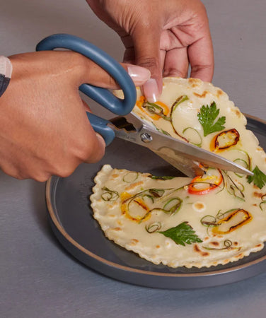 A person uses blue-handled scissors to cut a savory pancake topped with vegetables and herbs on a gray plate.