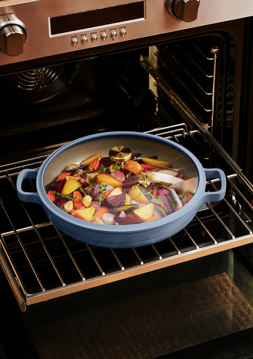 A blue roasting pan filled with chopped vegetables and herbs sits on an oven rack inside a stainless steel oven.
