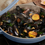 A blue pan filled with steamed mussels, charred lemon halves, and herbs, with steam rising from the food.