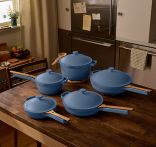A set of four blue cookware pots and pans with wooden handles are arranged on a wooden kitchen table in a modern kitchen setting.