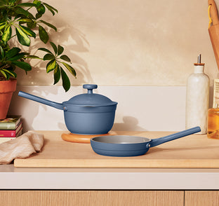 A blue saucepan with lid and a blue frying pan sit on a wooden kitchen counter next to a plant, oil bottle, and books.