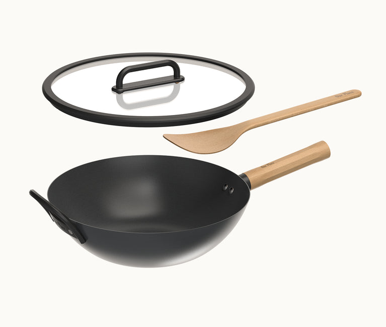 Black wok with wooden handle, glass lid, and wooden spatula on a white background