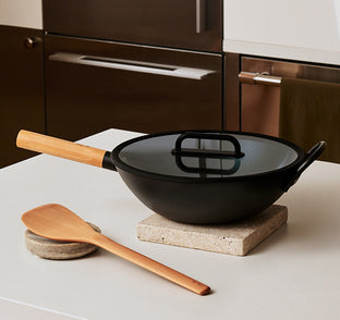 A black wok with a wooden handle and glass lid sits on a stone trivet; a wooden spatula rests nearby on a white kitchen countertop.