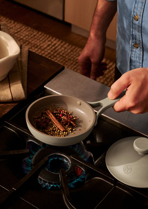 A person toasts spices in a small gray pan over a gas stove burner, holding the handle while wearing a light blue shirt.