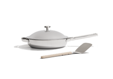 A stainless steel frying pan with a lid sits next to a metal spatula with a light-colored handle on a white background.