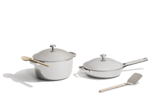 A white pot with lid and spoon, a white skillet with lid, and a spatula are arranged on a white background.