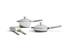 Two stainless steel saucepans with lids and long handles are placed next to a wooden spoon and a spatula on a white background.