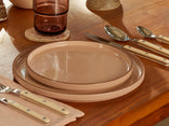 A set table with beige ceramic plates, cream-handled cutlery, a mauve glass on a stone coaster, and a folded napkin on a wooden surface.