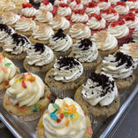 A tray of assorted cupcakes topped with various frostings and garnishes, including cookie crumbs, fruit pieces, and sprinkles, displayed in rows.