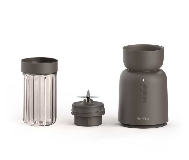 A compact blender set with three parts: a clear ribbed blending cup, a blade attachment, and a matte gray motor base with buttons and the brand name Our Place on the front, all on a white background.