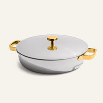 The Titanium Braiser Pro 11.5” is a round chef-approved stainless steel braiser with a lid, featuring gold-colored handles and a matching gold knob, set against a plain light background.