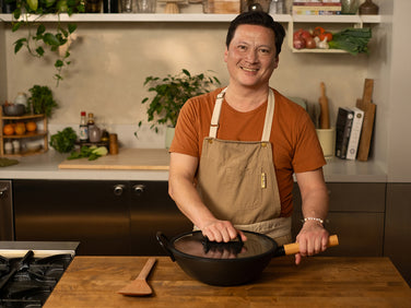 Man in a kitchen holding a wok with a lid, wearing an apron.