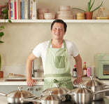 Man in a kitchen wearing a green apron with pots and pans in front of him.