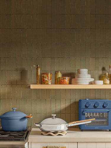 A kitchen scene with a blue enameled cast-iron pot on a stovetop, a white pan with a gold handle on a burner grate, and a blue toaster oven against a green tiled backsplash.