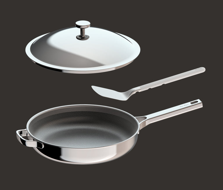 A stainless steel frying pan with a long handle, a matching lid shown above it, and a metal spatula placed beside the pan, all set against a plain dark background.