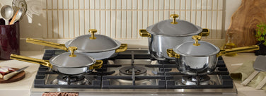 Four stainless steel pots and pans with gold handles are placed on a gas stove in a modern kitchen. Kitchen utensils, cutting boards, herbs, and garlic are visible on the countertop.