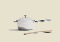 A white saucepan with a lid and metal handle sits on a light surface. In front of it lies a light wooden spoon. The background is a plain, off-white color.