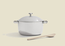 A shiny stainless steel pot with a lid and side handles is placed on a light surface, with a light-colored wooden spoon lying in front of it. The background is a solid beige color.
