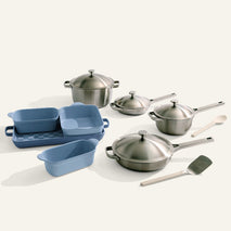 The Titanium Pro Cookware + Bakeware Set includes stainless steel and titanium pots with lids, two blue nonstick bakeware dishes, a wooden spoon, and a metal spatula arranged on a light surface.