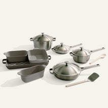 The Titanium Pro Cookware + Bakeware Set includes stainless steel pots with lids, pans, nonstick baking dishes and loaf pan, a wooden spoon, and metal spatula, all neatly displayed on a white surface.
