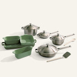 The Titanium Pro Cookware + Bakeware Set includes stainless steel pots and pans with lids, two green nonstick bakeware dishes, a green loaf pan, a wooden spoon, and a metal spatula displayed on a light surface.