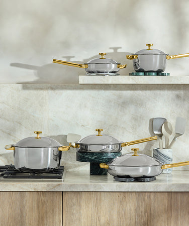 A set of shiny stainless steel pots and pans with gold handles is arranged on a kitchen counter next to a stovetop, white utensil holder, and small potted plant. The background features a light stone backsplash.