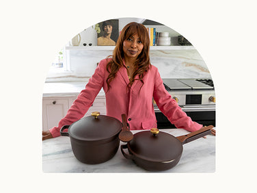 A woman in a pink shirt stands in a bright kitchen behind a marble counter with two dark brown cookware pieces, a saucepan and a skillet, placed in front of her.