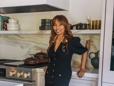 A woman with long brown hair in a black dress stands smiling in a modern kitchen, leaning against a marble countertop near a stove with brown cookware and neatly stacked dishes on open shelves.