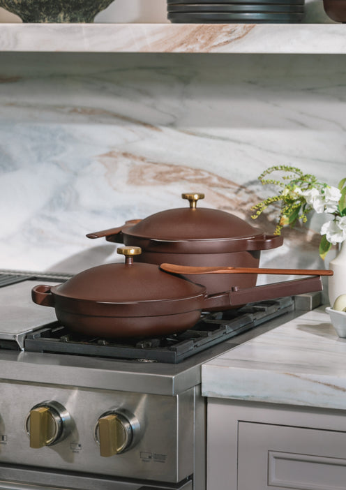 Two brown, lidded pans with gold handles sit on a stainless steel gas stove in a marble-accented kitchen, with a white vase containing green and white flowers nearby.
