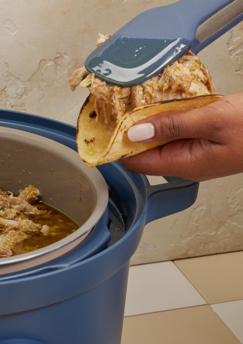 A hand holds a taco shell while using a spatula to fill it with shredded meat from a blue slow cooker on a tiled kitchen counter.