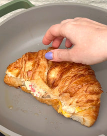 A hand with lavender nail polish holds a croissant filled with melted cheese and other ingredients inside a nonstick frying pan.