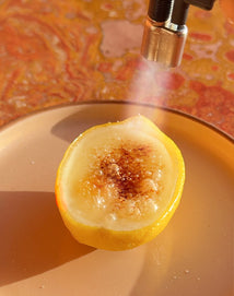 A lemon half on a beige plate is being caramelized on top with a kitchen torch, creating a bruleed surface. The background has a warm, orange and yellow marbled pattern.