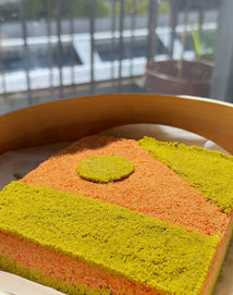 A square-shaped layered dessert with vibrant orange and green crumbly textures, topped with a round green piece, sits in a wooden steamer basket near a sunlit window.