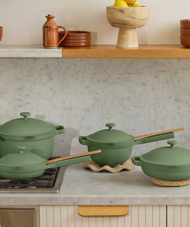 Four matte green pots and pans with wooden handles are arranged on a marble countertop; two sit on trivets, one is on a stove, and a beige vase with green foliage is in the background.