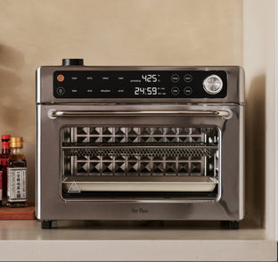 A stainless steel countertop oven with a digital display set to 425°F and 24:59 minutes, surrounded by control buttons and a knob, sits on a kitchen shelf next to bottles of oil and sauces.