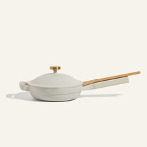 A modern off-white ceramic frying pan with a matching lid, wooden handle, and gold knob, shown against a plain light background.