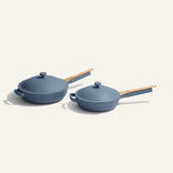 Two modern blue nonstick pans with matching lids and wooden handles are placed side by side on a light background. One pan is slightly larger and has an extra helper handle.
