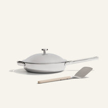The Titanium Always Pan Pro, a white lidded frying pan with a long handle and innovative nonstick technology, sits next to a metal spatula with a beige handle on a light background.