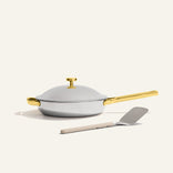 The Titanium Always Pan Pro, a white no-coating nonstick frying pan with a gold handle, sits on a light surface with a metal spatula placed in front.