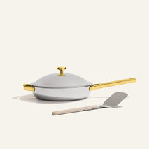 The Titanium Always Pan Pro, a white no-coating nonstick frying pan with a gold handle, sits on a light surface with a metal spatula placed in front.