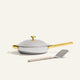 The Titanium Always Pan Pro, a white no-coating nonstick frying pan with a gold handle, sits on a light surface with a metal spatula placed in front.