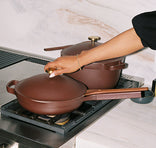 A person wearing a gold bracelet lifts the lid of a brown pan on a gas stove, with a matching brown pot in the background. Both cookware pieces have gold handles and the kitchen has a marble backsplash.