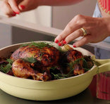 Person seasoning roasted chicken in a yellow pan with herbs